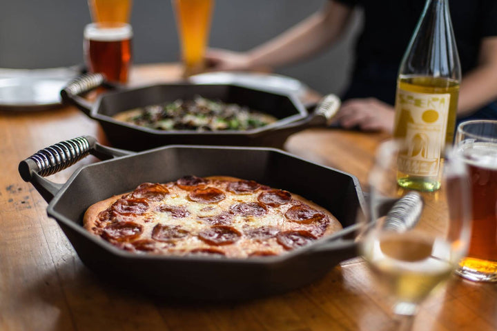 FINEX cast iron skillet with pepperoni pizza, surrounded by drinks on a wooden table, showcasing its unique octagonal design.