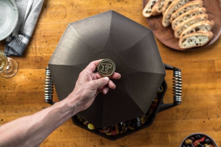 Hand lifting lid of FINEX cast iron skillet on wooden table with bread slices in the background.